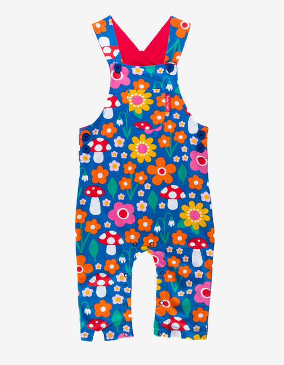 blue dungarees with a multicoloured flower and toadstool print, with navy poppers on straps and sides, a pocket on the front and red lining